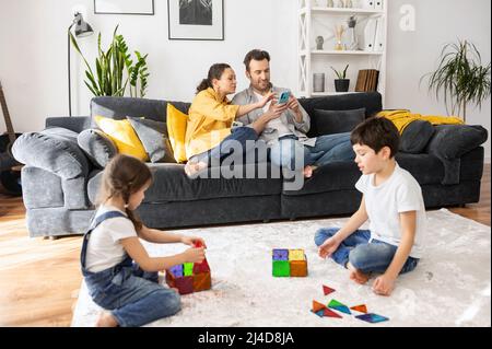 Zwei Eltern mit Smartphones sitzen auf dem Sofa im gemütlichen Wohnzimmer, scrollend Nachrichten-Feed, während zwei Kinder spielen mit Konstruktor auf dem Boden, Geschwister genießen Spiel zusammen. Vierköpfige Familie zu Hause Stockfoto