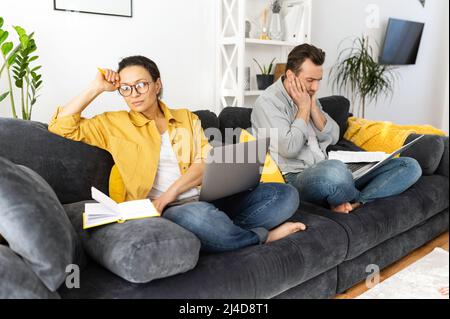 Das Paar streitet zu Hause, eine frustrierte Frau sitzt mit einem Laptop, ein trauriger Mann auf dem Hintergrund. Neuvermählte haben Schwierigkeiten, Beziehungskrise Stockfoto