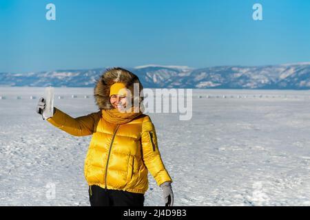 Glücklich lächelnde Frau trägt Winter gelbe Jacke hält Stück transparente zerkleinerte Eiswürfel über ihr Gesicht. Die Sonne scheint durch die Seiten des Eises Stockfoto