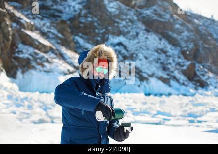 Mann in blauer Jacke gießt kochendes Wasser oder Tee aus einer Thermoskanne und trinkt an einem sonnigen Wintertag in einem starken Frost. Baikalinsel Stockfoto