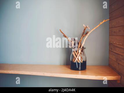 Neutrale farbige Vase mit Holzstäben und natürlichen Kegeln auf verzweifeltem Holzregal gegen raue hellgrau-blaue Wand im Sonnenlicht. Inneneinrichtung. Stockfoto