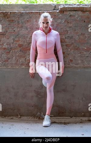 Junge schöne blonde Sportmädchen in rosa Sportswear. Die Athletin hält ihre Hände nach dem Training neben ihrem posierten Backsteinwand-Hintergrund. Tageslicht. Modernes Konzept. . Hochwertige Fotos Stockfoto