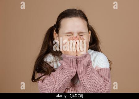 Porträt eines jungen ehrfürchtigen Mädchens mit langen dunklen Haaren in weiß, rosa Pullover stehen mit geschlossenen Augen, verschrauben Augen, den Mund mit Händen bedeckt Stockfoto