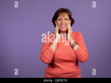 Frau mittleren Alters mit Kiefergelenkschmerzen, die ihren Kiefer halten und einen schmerzhaften Ausdruck machen. Vorstellung des Konzepts „chronische Schmerzen“. Stockfoto