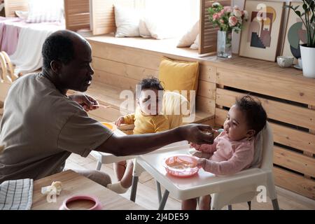 Moderne liebende afroamerikanische Vater verbringt Zeit mit Baby Töchter Fütterung sie mit Fruchtpüree Stockfoto
