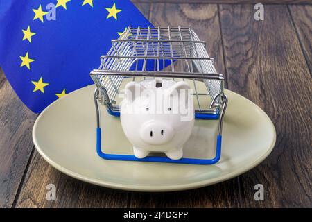 Sparschwein auf einem Teller unter dem Warenkorb und der Flagge der Europäischen Union. Konzept zur Inflation von Lebensmitteln Stockfoto