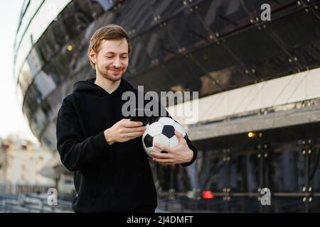 Lächelnder Fußballfan mit Fußball, der Smartphone hält und die Wetten-App nutzt. Fußballstadion im Hintergrund. Speicherplatz kopieren. Spielkonzept. Stockfoto