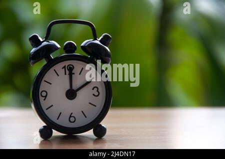 Schwarzer Wecker, isoliert auf unscharfem Naturhintergrund. Die Uhr wurde auf 2 Uhr eingestellt. Speicherplatz kopieren. Stockfoto