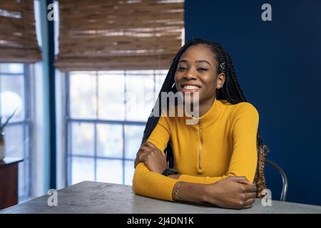 Junge afrikanische amerikanische Frau mit gelben Hemd an der Arbeitsplatte in der Küche der Insel sitzen. Trendy ethnische lifestyle Bild. Stockfoto