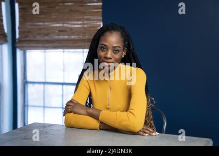Junge afrikanische amerikanische Frau mit gelben Hemd an der Arbeitsplatte in der Küche der Insel sitzen. Trendy ethnische lifestyle Bild. Stockfoto