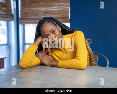 Junge afrikanische amerikanische Frau mit gelben Hemd an der Arbeitsplatte in der Küche der Insel sitzen. Trendy ethnische lifestyle Bild. Stockfoto