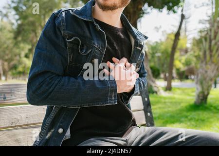 Herzschmerz, der junge Mann hat einen Herzinfarkt, Herzleiden des jungen Mannes, Erwachsener sitzt auf Parkbank und hält die Brust Stockfoto