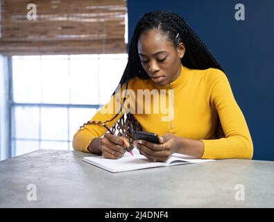 Junge afrikanische amerikanische Schüler mit gelben Hemd an der Theke Insel Schreiben in ein Notebook zu sitzen. Trendy ethnische lifestyle Bild. Stockfoto