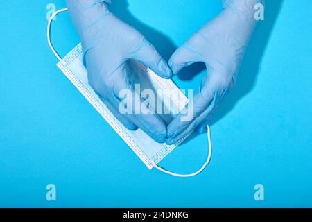 Hände in Latexhandschuhen machen Zeichen in Form von Herz auf dem Hintergrund der blauen medizinischen Maske close-up Stockfoto