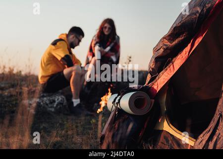 Rucksack mit Matte und Zelt im Fokus, auf dem Hintergrund zwei junge Reisende Mann und Frau sitzen beim Sonnenuntergang am Lagerfeuer, Reisekonzept Stockfoto