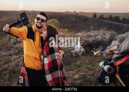 Junges Paar, das am Abend Selfie auf dem Gipfel des Hügels vor dem Lagerfeuer und Zelt macht Stockfoto