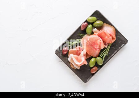 Draufsicht auf Charcuterie-Tafel mit Proscuitto serviert mit Oliven auf weißem Steintisch Hintergrund mit Kopierplatz. Schwarzer Teller mit traditionellem italienischen Stil Stockfoto