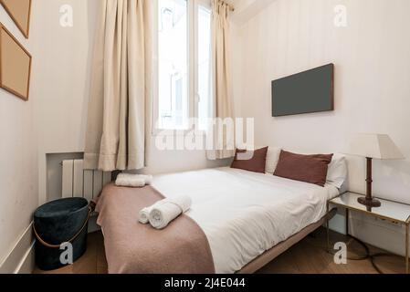 Schlafzimmer mit großem Bett mit weißen Laken, braunen Kissen, Aluminiumheizkörper, Beistelltisch aus Eisen und Glas und braunem Parkettboden Stockfoto