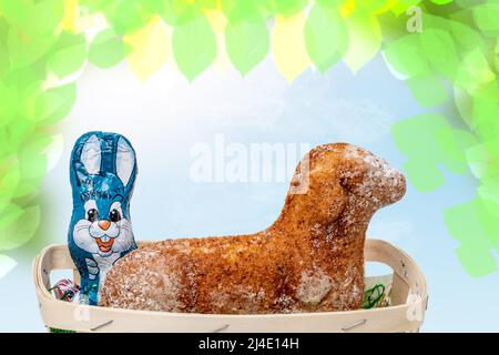 Vorlage für die Osterkarte. Ein traditioneller Oster-Lammkuchen und ein Osterhase und viele andere Schokoladenbonbons in einem Korb über abstraktem Frühlingshintergrund Stockfoto