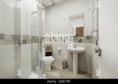 Beigefarbenes Badezimmer mit dunklem Volance und rahmenlosem Wandspiegel, weißem Porzellanwaschbecken mit passendem Sockel, weißem Handtuchheizkörper und Chromakzent Stockfoto