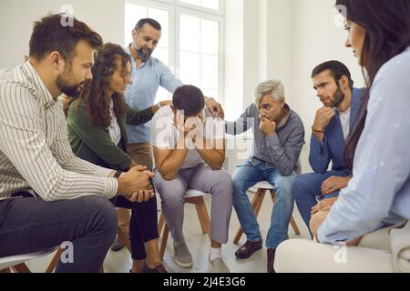 Menschen, die einen weinenden jungen Mann während einer Gruppentherapie unterstützen und trösten Stockfoto