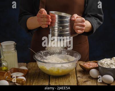 Nahaufnahme. Ein Koch in dunkler Uniform siebt Mehl durch ein Sieb auf einem hölzernen Küchentisch. Mehl im gefrorenen Flug. Herstellung von Teigprodukten - Stockfoto