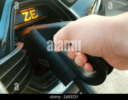 Ein Elektroauto von Renault Zoe, das mit dem schnellen EV-Ladestecker aufgeladen wird, der zum Laden durch die Hand einer Person in die Autosteckdose gesteckt wird. Stockfoto