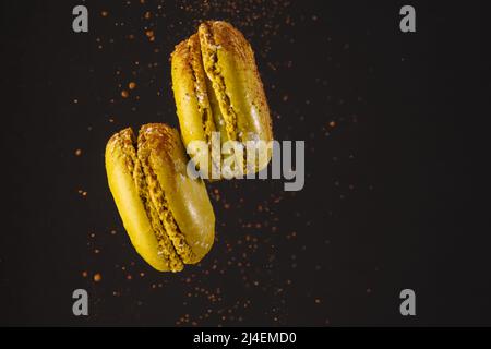 Gelbe Makronen: Freifliegendes französisches Gebäck auf schwarzem Hintergrund mit Schokoladenbröseln. Köstliche süße kalorienreiche Speisen, festliche Leckerbissen. Es gibt f Stockfoto