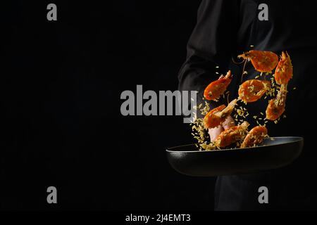 Ein professioneller Koch in schwarzer Uniform bereitet in einer Bratpfanne auf schwarzem Hintergrund Garnelen zu. Gefrorenes Essen während des Fluges. Restaurant, Hotel, Rezept bo Stockfoto