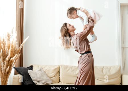 Lächelnde Mutter hebt die glückliche kleine Tochter im Wohnzimmer in die Luft und spielt mit ihr Stockfoto