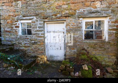 Ein altes Haus in Langthwaite, Archengarthdale, Yorkshire Dales National Park. Die Farbe blätterte von der alten Holztür und den Fenstern in einer alten Steinwand ab. Der Stockfoto