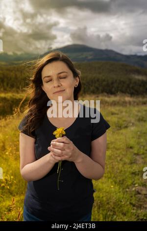Schöne hispanische Frau mit geschlossenen Augen, die in der Mitte des Feldes mit Blick auf die Kamera steht und gelbe Blumen riecht, die sie in sich hält Stockfoto