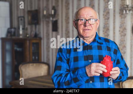 Älterer europäischer kahler älterer Mann in blauem Flanellhemd mit künstlicher gefälschter Herzstütze vor seiner Brust. Altmodische Innenausstattung im Hintergrund. Gesundheitskonzept. Hochwertige Fotos Stockfoto