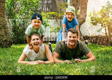 Glücklich lächelnde Familie mit zwei Kindern liegen im Hause Hinterhof Garten Gras. Blick auf die Kamera Stockfoto