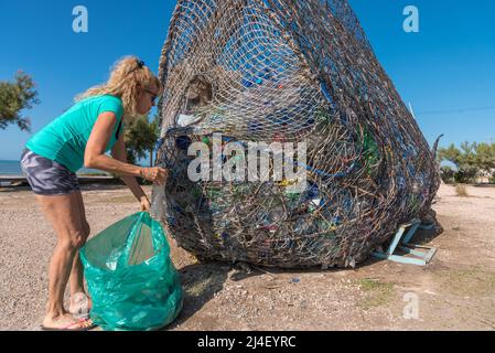 Reife blonde Frau mit Sonnenbrille, die eine Plastikflasche in einen Abfalleimer voller recycelbarem Material legt Stockfoto