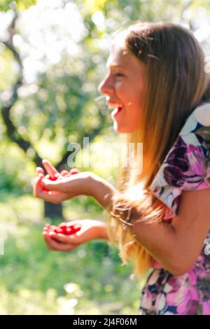 Defocus schöne lächelnde Teenager-Mädchen im Kleid lächelt vor grünen Sommer Hintergrund. Abstrakter Sommerhintergrund. Teen Mädchen hält Himbeeren fru Stockfoto