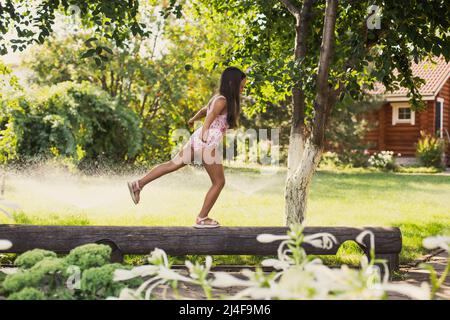 Das weibliche Kind balanciert auf einem Fuß auf dem Holzbaum und sieht aus wie eine Schwalbe, die tagsüber mit Wassersprinklern, Haus und grünen Bäumen im Hintergrund verwöhnt wird. Träumen Stockfoto