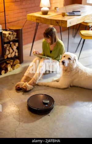 Der Roboter-Staubsauger arbeitet auf dem Boden, während die Frau mit einem Hund zu Hause am Kamin sitzt Stockfoto