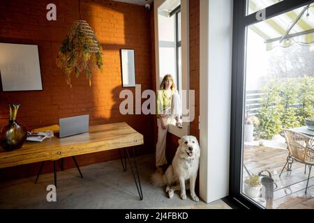 Frau, die auf der Fensterbank sitzt und mit ihrem weißen, entzückenden Hund zu Hause aus dem Fenster schaut Stockfoto