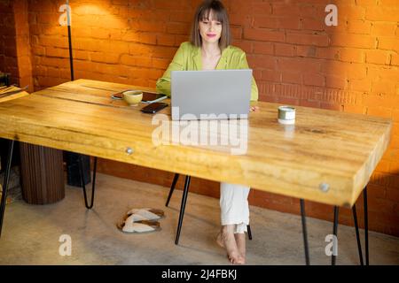 Die Frau arbeitet am Laptop, während sie am Holztisch im gemütlichen Zuhause sitzt Stockfoto