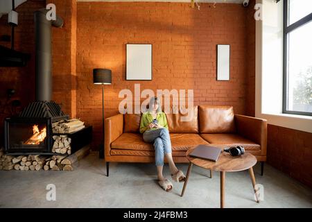 Frau sitzt mit Gadgets im modernen Wohnzimmer im Loft-Stil zu Hause Stockfoto