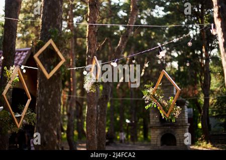 Hochzeitsfeier im Freien im Pinienwald. Esstische, Bänke im Boho-Stil mit Kerzen, Blumen, Frühlings-Lichterketten, Teller dekoriert Stockfoto