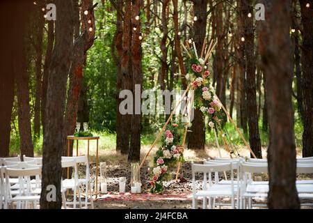 Wunderschöne böhmische Tipi-Bogendekoration auf der Hochzeitsfeier im Freien im Pinienwald mit Kegeln. Stühle, floristische Blumenkompositionen aus Rosen Stockfoto