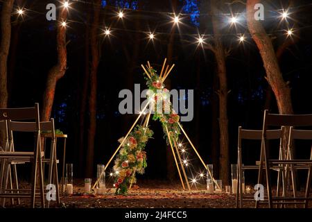 Festliche Beleuchtung mit Fadenlampen auf Boho Tipi-Bogen am Veranstaltungsort für Hochzeitsfeiern im Freien im Pinienwald bei Nacht. Glühbirne mit Vintage-Schnur Stockfoto