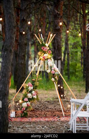 Böhmischer Tipi-Bogen aus Holzstäben, verziert mit rosa Rosen, Kerzen auf dem Teppich, Pampass-Gras, eingewickelt in Lichterketten auf der Hochzeit im Freien Stockfoto