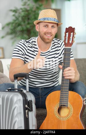 Männlicher Musiker mit Koffer und Gitarre, der die Daumen hoch hält Stockfoto