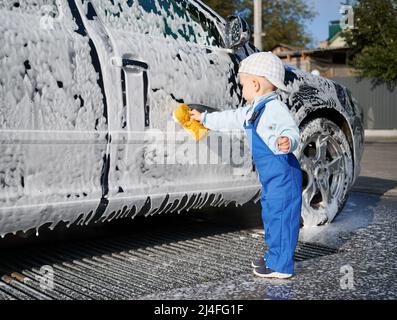 Kleines männliches Kind in blauem Jumpsuit, das versucht, zu studieren, wie man Auto waschen kann, bedeckt mit weißem Schaum mit Hilfe eines gelben Schwamms. Kleinkind Junge lernen neue Lektion der Autoreinigung. Stockfoto