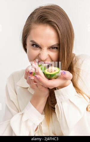 Porträt einer reizenden, lächelnden jungen, brünette kaukasischen Frau, die das weiße Hemd mit langen Haaren trägt und Avocado zeigt, isoliert auf weißem Hintergrund stehend Stockfoto