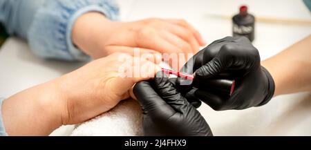 Maniküre Lackierung. Nahaufnahme eines Maniküre-Meisters mit schwarzen Gummihandschuhen, die roten Lack auf einen weiblichen Fingernagel im Schönheitssalon auftragen Stockfoto