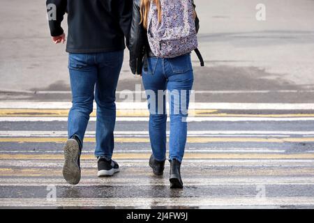 Ein Paar in Jeans geht auf Fußgängerübergängen und hält sich die Hände. Straßenzebra-Markierung, Menschen auf nassem Fußgängerüberweg im Frühlingswetter, Straßensicherheitskonzept Stockfoto